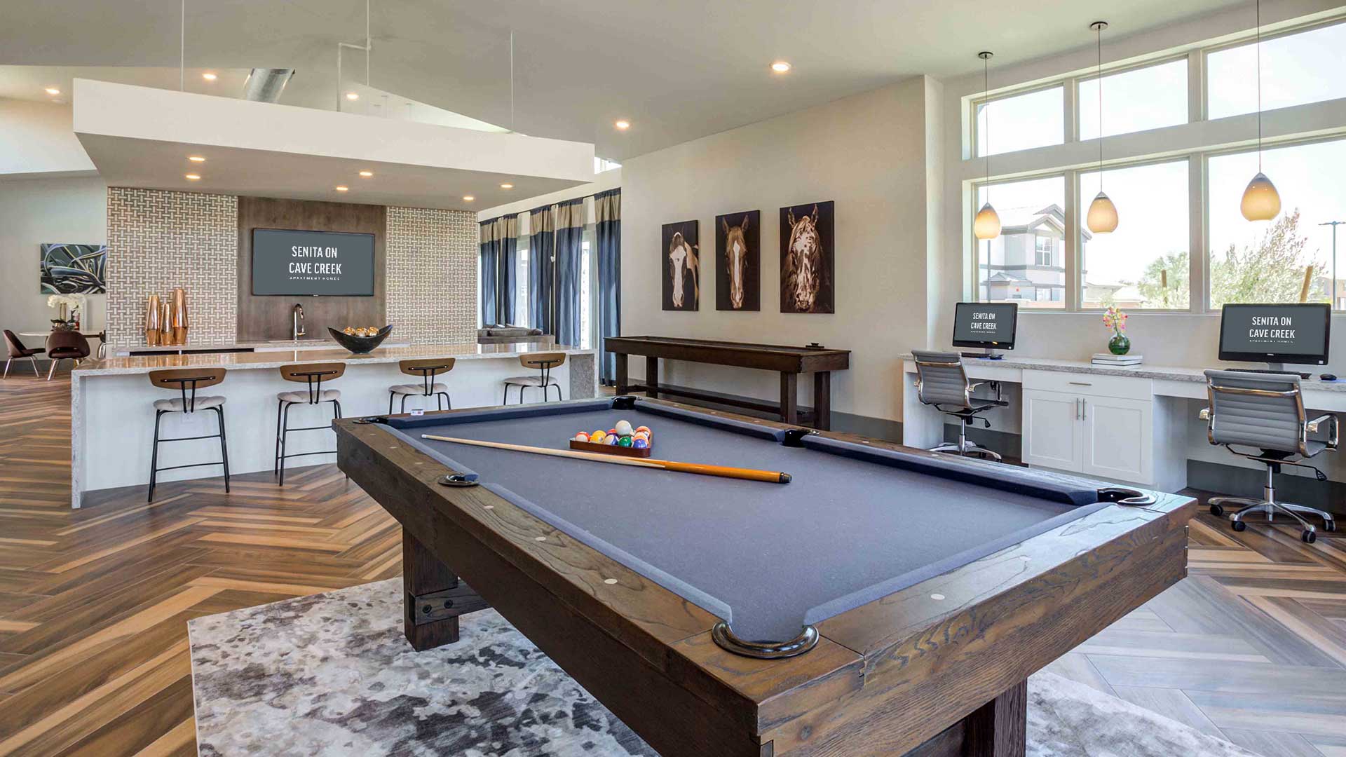 Modern home interior with a pool table, kitchen bar stools, and a home office setup, featuring large windows and hardwood flooring.