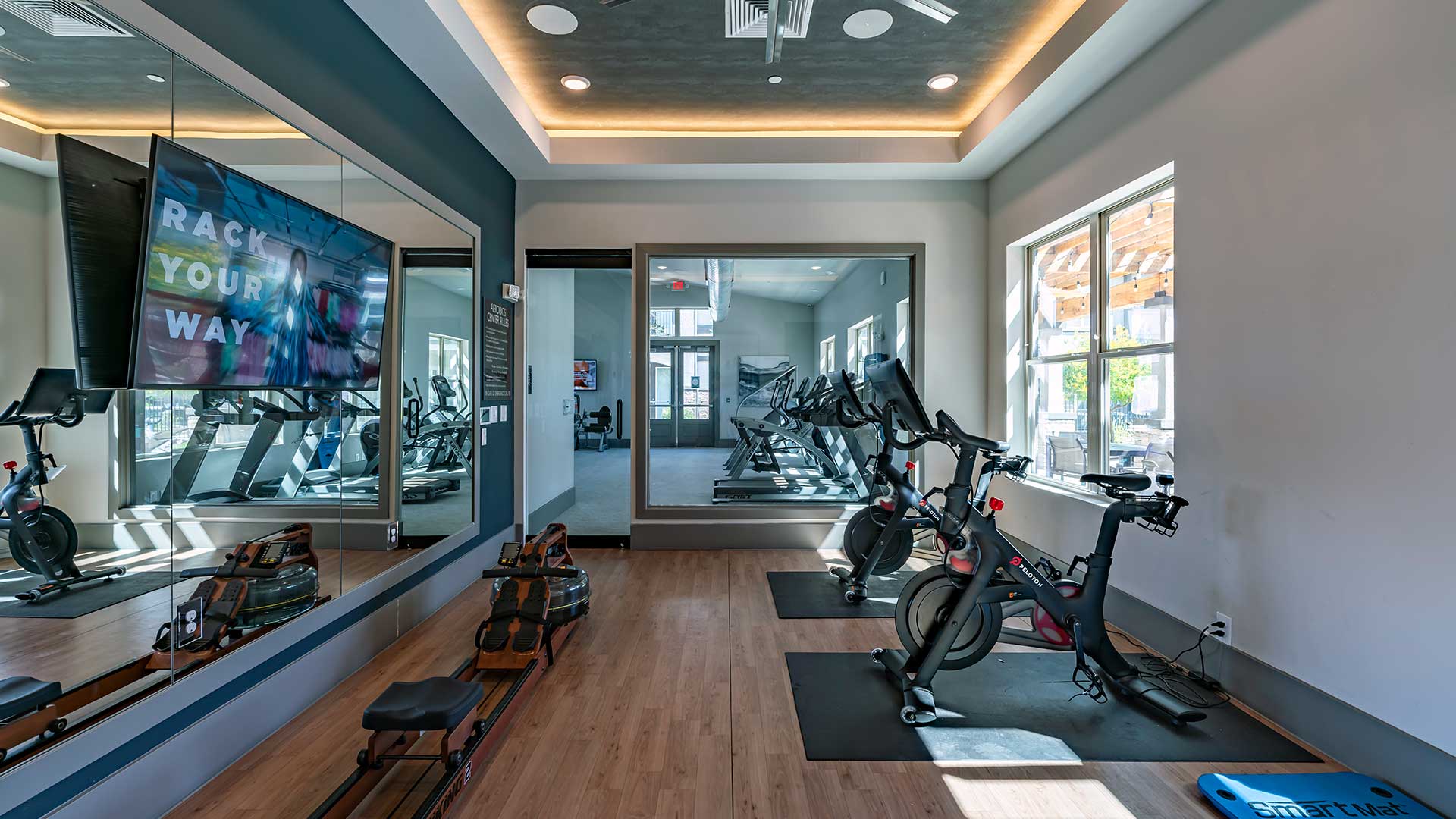 Interior of a modern gym with stationary bikes, treadmills, and a mirrored wall, featuring a large screen displaying motivational text.