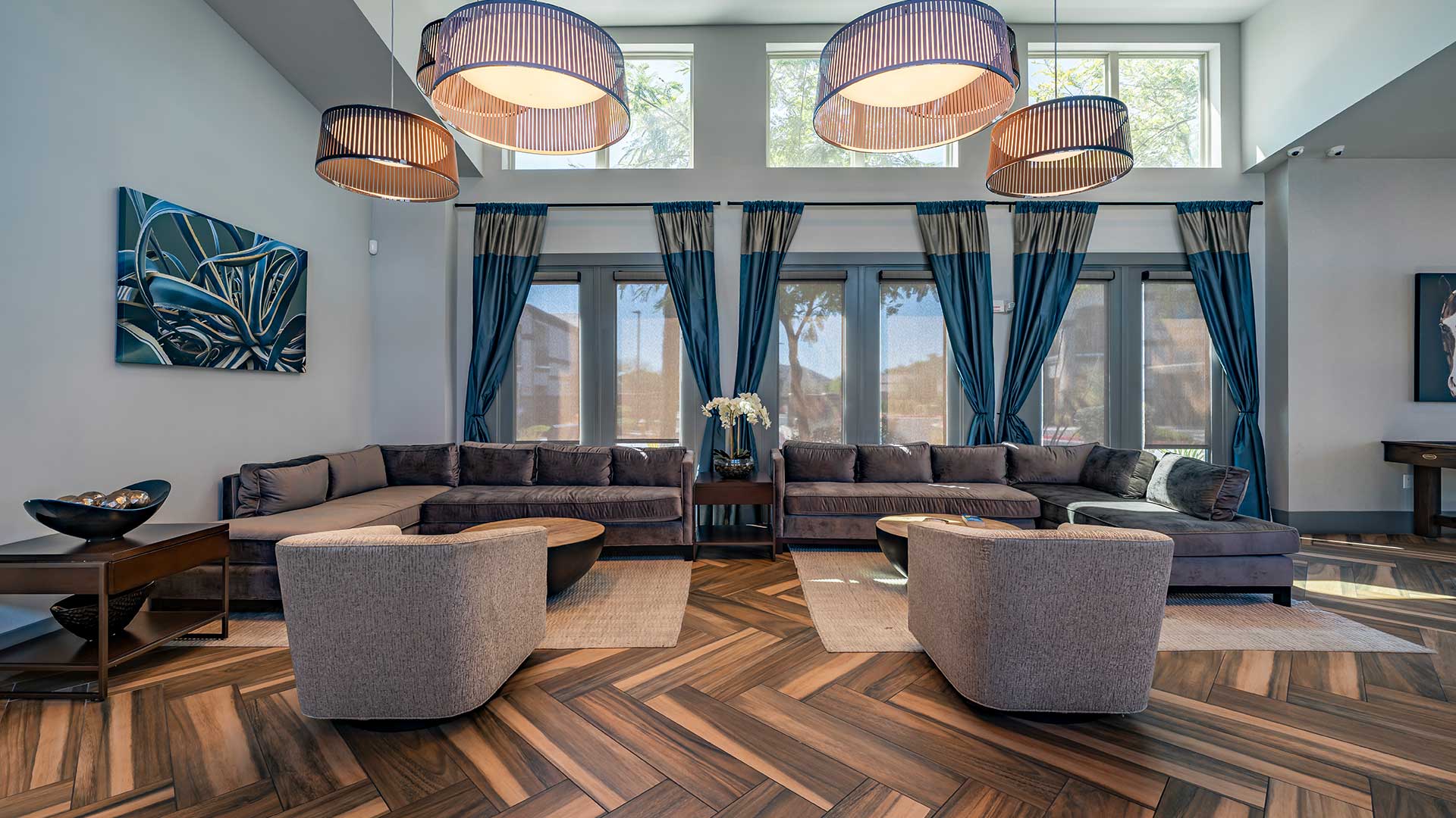 Modern living room featuring dark brown sofas, gray armchairs, wooden herringbone floor, large windows with curtains, and contemporary light fixtures.