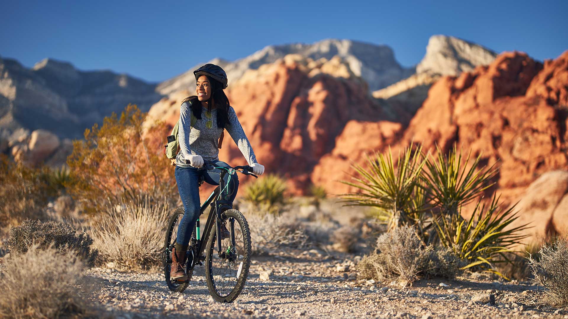 A woman cycling on a desert trail with red rock formations in the background, wearing a hat and backpack, during the daytime.