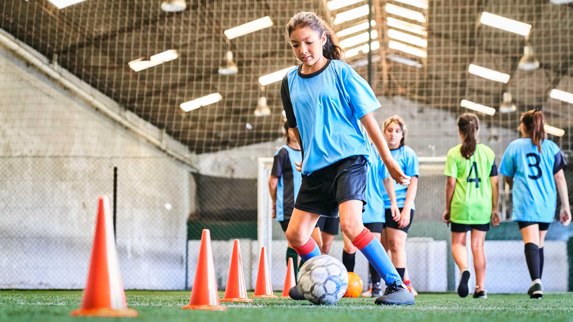 A young girl in a blue jersey and red socks dribbles a soccer ball through orange cones on an indoor field, with other players in the background.