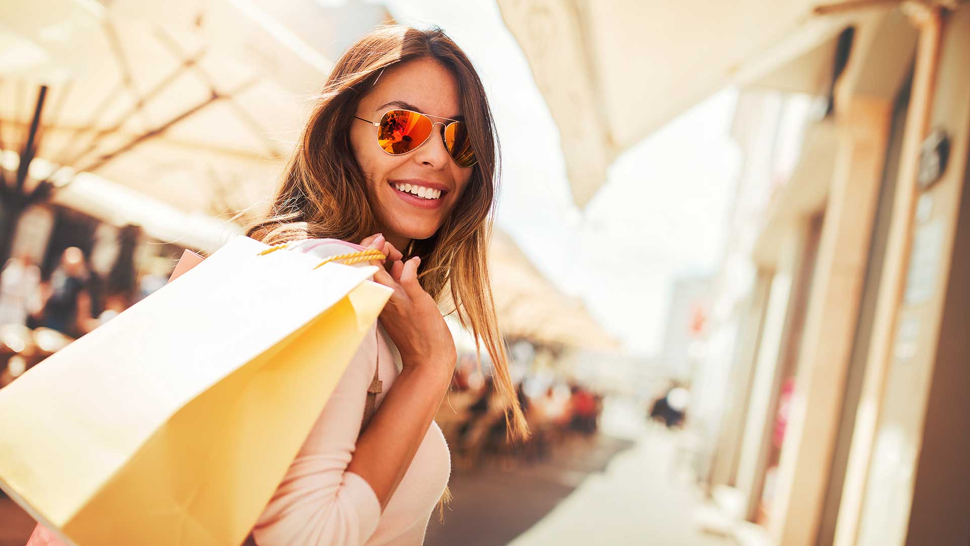 A cheerful woman in sunglasses holding shopping bags on a sunny street, reflecting a lively urban shopping atmosphere.