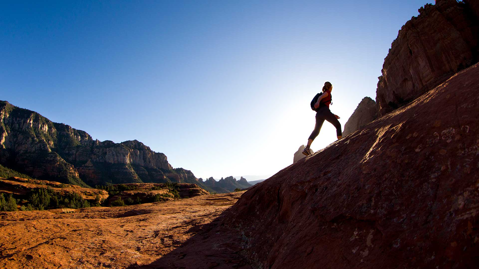 A hiker with a backpack ascends a steep, rocky trail with majestic mountains under a clear blue sky in the background.