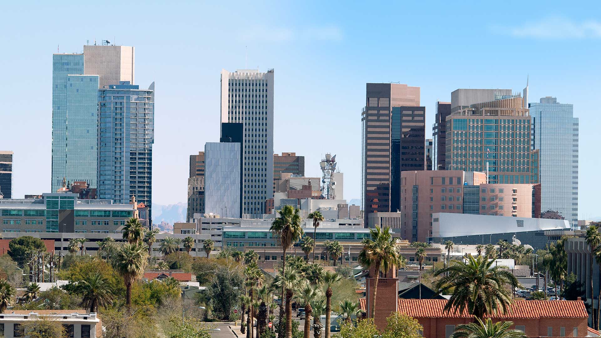 Skyline of phoenix, arizona, featuring modern high-rise buildings under a clear blue sky with scattered palm trees in the foreground.