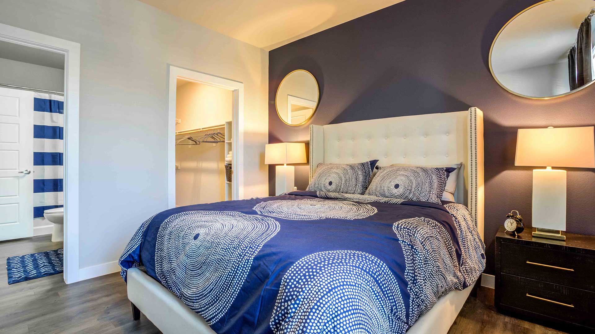 Modern bedroom with a white tufted headboard, navy blue bedding, round mirrors on a dark blue wall, and wooden nightstands with lamps.