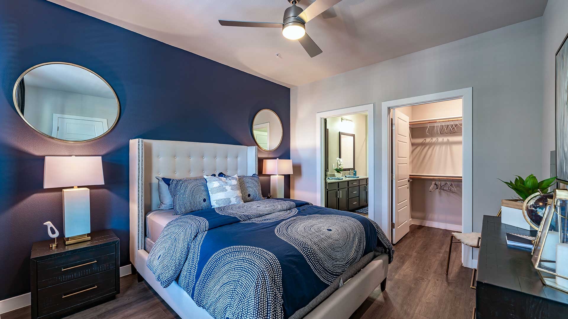 Modern bedroom with a navy and white color scheme, featuring a plush bed, circular mirror, bedside lamps, and an open closet.